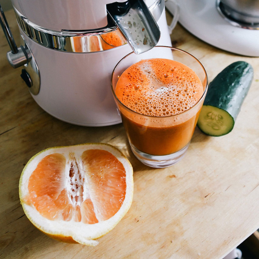a juicer pours orange juice into a cup. half of an orange and a cucumber is on the table